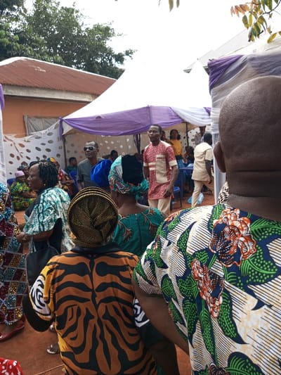 The scene depicts an outdoor social gathering in Oko Town, Nigeria. The foreground shows the backs of three individuals: on the right, a person with a shaved head wearing a patterned shirt featuring green leaves, white stripes, and red-orange flowers on a dark blue background; in the center, a person wearing a brown and black tiger-stripe patterned top and a woven, straw-colored head covering; and on the left, a woman with dark hair wearing a patterned light green and white top.
In the midground, under a large white canopy draped with purple fabric accents, multiple individuals are gathered. A man in a light red and beige patterned traditional shirt and light trousers stands in the center, looking right. Behind him, to the right, another man in a light-colored traditional outfit walks away from the viewer. To the left, a man wearing dark sunglasses and a dark grey shirt looks towards the right. Other attendees, including women in various patterned Nigerian attire and a woman in a yellow top, are visible further under the canopy, some seated on blue plastic chairs.
The background includes a building with an orange-brown wall and a reddish-brown corrugated metal roof on the left, beside the canopy. White fabric with large polka dots serves as a decorative or privacy screen on the left side of the canopy. The ground is reddish soil. Green trees and foliage are visible against a light sky above and behind the structures, with some branches in the upper right corner.