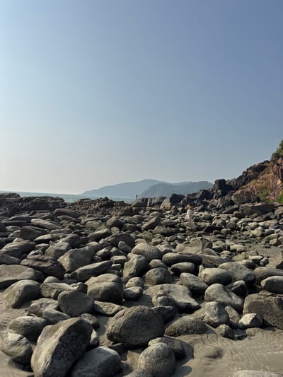 A landscape view shows a mass of rounded, gray rocks stretching along a beach, with some patches of sand visible between them. A mountain range is faintly visible in the background, partially obscured by haze. A cliff covered with greenery is at the top of the image. The sky is clear and light blue. A small figure in white stands among the rocks.