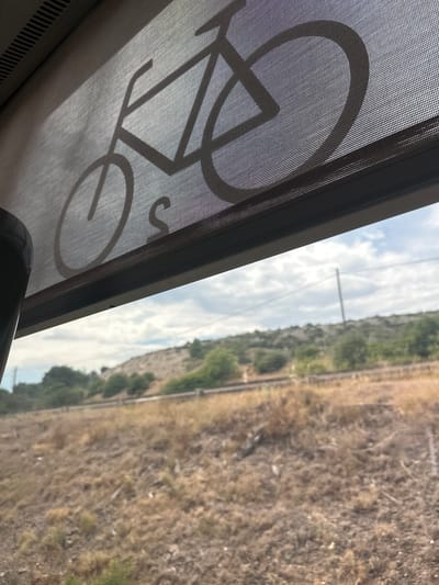The image captures a view from inside a moving vehicle, likely a train, looking out of a window. A mesh screen with a silhouette of a bicycle is visible in the foreground, positioned across the bottom left of the frame. The bicycle graphic appears to be a symbol indicating bicycle transport. Beyond the screen, the window reveals a landscape dominated by dry, scrubby vegetation and a clear sky with scattered clouds, suggesting a sunny day. A utility pole is visible in the distance, and a faint track runs through the landscape, hinting at the train's path. The overall impression is of travel through a rural or semi-arid environment. No people are visible, and there is no discernible text in the image. The lighting suggests it is daytime, possibly late morning or afternoon. The location context provided is Salses-le-Château, France, which aligns with the arid appearance of the landscape.