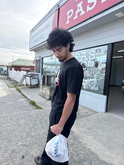 A young adult male stands on a concrete sidewalk in Tolhuin, Argentina, facing left with his head tilted downward. He has dark, curly hair, wears clear-framed glasses, and a black t-shirt with a partially obscured graphic, black pants, and dark sneakers. A silver bracelet is visible on his right wrist. He holds a white plastic bag labeled "LA LYON PANADERÍA CONFITERÍA" in his right hand. The individual is positioned in front of a commercial building with a white facade and red signage that reads "TRADICIÓN FUEGUINA DESDE 1932" and "PAS." The storefront features large glass windows displaying an outlined drawing of a cow's head and interior elements. To the left of the individual, a white picket fence, a metal refuse container, and several modest wooden buildings with light-colored siding and red roofs are visible further down the street. The sky is overcast.