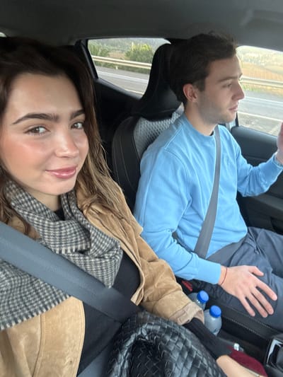 The image depicts two individuals inside a moving vehicle, presented as occurring within or near Mafra, Portugal. A female, positioned in the front left of the frame (likely the passenger seat from her angle or a selfie from the driver's side, though no steering wheel is visible), faces the camera directly, holding a device to capture a selfie. She wears a light brown suede jacket over a black top, with a grey and white plaid scarf around her neck. Her hair is brown with lighter highlights, and she has visible makeup. A grey seatbelt crosses her chest and lap. To her right, a male sits in what appears to be the front passenger seat. He wears a light blue crew-neck sweatshirt and dark blue or grey trousers. A grey seatbelt is visible across his chest. He is oriented towards the right of the frame, looking forward and slightly upward, with his left hand raised and partially open, gesturing or resting. Two clear plastic water bottles with blue caps are visible in the center console area between the two individuals. A black woven bag rests on the lap area of the female. Through the car window on the right, a multi-lane road with a white guardrail is visible, alongside vegetated hills under an overcast or late afternoon sky.