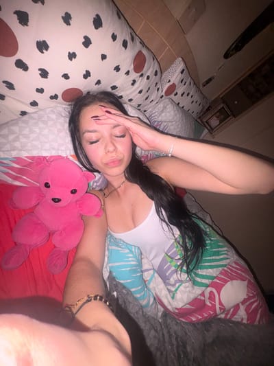 A young woman lies on a bed with patterned bedding. She wears a white top and has long, dark hair. Her eyes are closed, and one hand covers her forehead while the other rests beside a pink stuffed octopus. She wears a bracelet on each wrist and a necklace. The bed is topped with patterned pillows and a gray blanket. A wooden headboard and wall shelf are partially visible in the background.