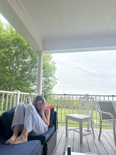 A young woman with long brown hair and glasses, wearing a blue tank top and striped pajama pants, sits cross-legged on a dark blue outdoor couch on a porch. She is looking down and touching her face. The porch has a white railing and a white roof supported by a white pillar. Beyond the railing, there is a green lawn, more white railings, and some buildings in the distance under a cloudy sky. The porch floor is made of grey composite decking. There are two white chairs and a grey lounge chair on the deck.