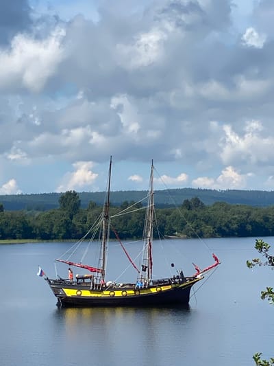 A traditional sailing vessel with a black hull and yellow accents is anchored on calm, dark blue water under a partly cloudy sky. The boat features two masts with rigging and red sails unfurled. Several people are visible on the deck of the boat. The background consists of a dense, dark green treeline with rolling hills beyond, suggesting a rural or natural landscape. The scene is likely set in or near Boyarshchina, Russia, given the context.