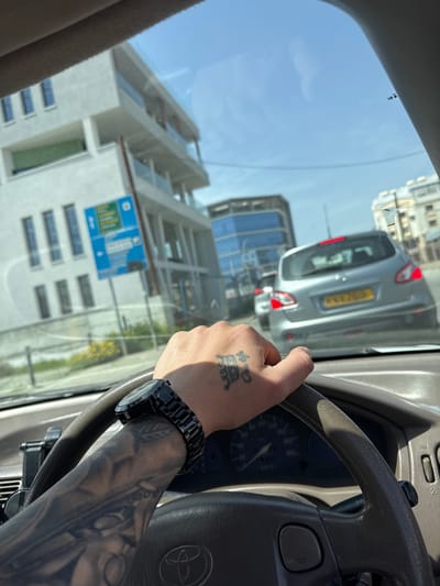 The scene is viewed from inside a vehicle, looking forward through the windshield in Kato Polemidia Municipality, Cyprus. A person's left arm and hand are prominently featured in the foreground, gripping a dark grey steering wheel. The arm displays extensive black-and-grey sleeve tattoos, while the back of the hand features a distinct black tattoo resembling an ornate letter or symbol. A black, chunky watch with a link bracelet is worn on the left wrist. The steering wheel prominently displays a Toyota logo in its center. A segment of the dashboard and speedometer, indicating a low speed, is visible behind the steering wheel, along with a black phone mount attached to the left dashboard vent.

The vehicle is either stopped or moving slowly in traffic. Directly ahead is the rear of a silver/light grey first-generation Nissan Qashqai. Its brake lights are illuminated, and the yellow Cypriot license plate, "KXX 785," is clearly visible. Another vehicle is partially visible further ahead.

To the left of the road, a modern, light-colored, multi-story building with vertical windows and glass-fronted balconies is present. In front of this building, a blue directional street sign with white text, likely in Greek, is positioned. Below the sign, a patch of ground with green vegetation and yellow flowers is visible. To the right, beyond the Nissan, other urban buildings are seen, including one with reflective blue glass elements. The sky above is clear blue with some light haze, indicating sunny weather.