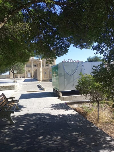 In Shar-Bakr, Uzbekistan, a street scene unfolds with a focus on a large, light-colored shipping container situated on a raised concrete platform. The container features two prominent blue hoses draped across its side. Above the container, the word "dijon" is visible in large, red lettering, likely part of a sign. To the left of the container, an ornate, light-colored building with arched windows and a domed turret suggests historical architecture. In the foreground, a wooden bench with concrete supports sits on a paved walkway. Overhead, the branches and leaves of trees cast dappled shadows on the walkway and the bench, indicating a sunny day. The area beyond the container appears to be a dry, grassy embankment.