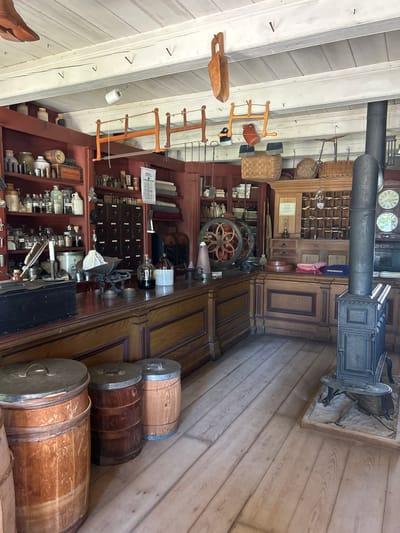 This media file shows the interior of a historical general store, likely in South Dundas, Canada. The scene is a cluttered and well-stocked shop, filled with an array of antique objects. Prominently featured is a dark metal wood-burning stove with a tall chimney pipe extending upwards, positioned in the upper right of the frame. Below it, the floor is made of light-colored wooden planks.

The walls and ceiling are constructed from white-painted wooden planks. Hanging from the ceiling are various wooden tools and implements, including what appear to be yarn winders and a wooden scoop. The shelves are packed with a diverse collection of items, such as jars containing liquids, rolled fabrics, boxes, and what might be apothecary bottles. A large, multi-drawer cabinet with a glass display case on top is visible in the center, possibly a post office or display cabinet. In front of this cabinet, a large, ornate industrial machine with two spoked wheels is present, suggesting a manufacturing or repair aspect to the store's former operations. Three wooden barrels with metal hoops are stacked next to each other along the right wall, adding to the rustic feel.

The overall impression is of a preserved historical space, showcasing the goods and equipment of a bygone era. There is no visible activity or event taking place, and no people are present. The lighting suggests it could be daytime, but the interior setting does not provide clear weather cues. Some visible text includes a "Post Office" sign within the glass display case. The scene evokes a sense of nostalgia and historical preservation.