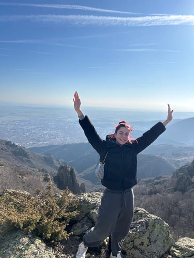 The image portrays a lone female individual positioned on a rocky mountain summit, overlooking a panoramic view identified as being near Ichera, Bulgaria. The subject, with red hair, wears a black puffer jacket, dark grey sweatpants, and white sneakers, carrying a light-colored, patterned shoulder bag. She smiles broadly, looking directly at the viewer, with both arms raised above her head, palms open, in a gesture of triumph or joy. The foreground consists of large, moss-covered rocks and dry, sparse shrubbery. The midground presents a deep valley with rugged, rocky terrain and dormant vegetation, likely trees without leaves. In the far distance, a sizable urban area or city sprawls across a hazy plain. The sky is clear blue, traversed by several faint contrails. The scene is brightly lit by natural sunlight.