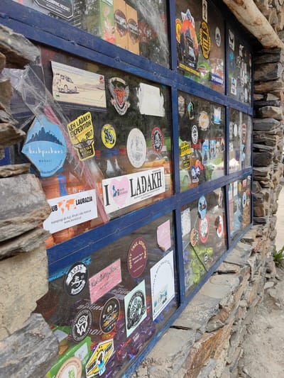 The image shows a shop window in Jispa, India, adorned with numerous stickers. The window is framed by a blue metal structure and surrounded by a stone wall. Inside the window, various products are visible, including bottles of beverages and packaged food items. The stickers, predominantly featuring travel and adventure themes, are applied to the glass, creating a layered collage. Some stickers advertise expeditions, motorcycle trips, and locations like Ladakh and Manali. A prominent sticker reads "LEH LADAKH EXPEDITION 2017." The overall scene depicts a commercial establishment catering to tourists and adventurers in a mountainous region.