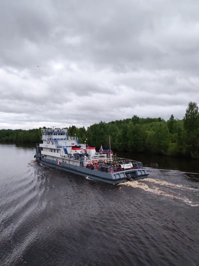 The image shows a riverboat traveling along a dark waterway in Srednii Rubezh, Russia. The boat is multi-tiered, colored primarily in gray, white, and blue, and labeled "OT-2446". Dense green vegetation lines both banks of the river. Overcast skies dominate the background. A small bird is visible in the sky. The boat creates a noticeable wake as it moves through the water.