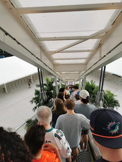 A low-angle shot shows a crowd of people on an escalator within a white, overhead structure. Some individuals wear Miami Dolphins apparel, including a hat. Palm trees and a row of gray stanchions are visible through the open sides. A person in a red and black outfit walks on the white pavement below, to the left of the moving walkway.