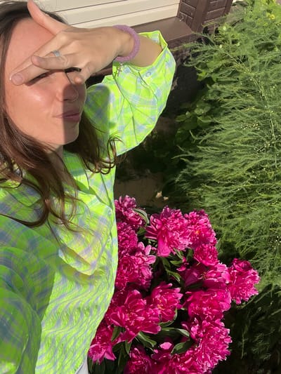 A young woman is taking a selfie outdoors, in front of some foliage and a house. She is wearing a bright green and blue checkered shirt and has her hand over her eyes to block out the sun. A ring is visible on her finger. To the right of the woman, a large bouquet of bright pink peonies is visible. The background shows green foliage, a portion of a house, and a textured surface that could be part of the house's exterior. The image appears to have been taken during the day, with strong sunlight casting shadows. This scene is located in Pigozino, Russia.