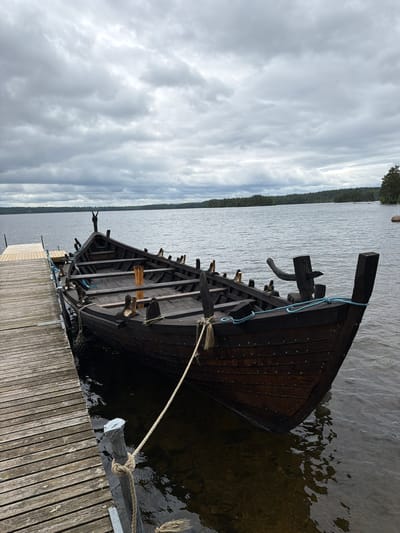 A dark wooden longboat is moored to a wooden pier on a calm lake under an overcast sky. The longboat has a pointed bow and stern and appears to be of historical design, possibly a replica. It is secured by ropes to posts on the pier. The water is dark and still, reflecting the cloudy sky. In the background, a tree-lined shore is visible across the lake. The scene suggests a quiet, natural setting in Aneby, Sweden.
