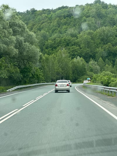 The image depicts a view from a moving vehicle on a two-lane asphalt road in Usakly, Russia. The road is bordered by a metal guardrail on the right and a dense, green forest on the left and in the background. The forest appears to be comprised of deciduous trees, with varying shades of green foliage. The sky is overcast, with a light gray hue.
There are several cars on the road. In the foreground, a silver sedan is visible from the rear, with its license plate reading "M914MA 702". Ahead of this car, two other vehicles are also traveling in the same direction. Further up the road, another white car is seen in the distance.
The road itself is in good condition, with clear white lane markings. The overall impression is of a scenic drive through a heavily wooded area.