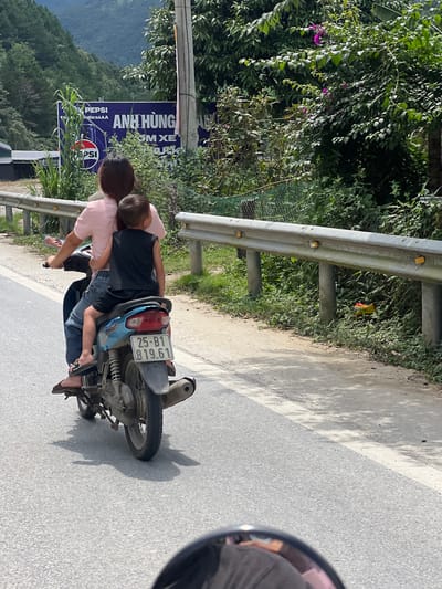 A woman and a young child ride on a motorcycle on a paved road. The woman is in the front, wearing a pink t-shirt and jeans, with dark hair. The child, wearing a black t-shirt and shorts, sits behind her, leaning his head on her shoulder. They are both wearing flip-flops. The motorcycle is blue and has a license plate that reads "25-B1 819.61".
The scene is outdoors, with lush green foliage on the left side of the road, indicating a mountainous or hilly area. A metal guardrail runs along the edge of the road. In the background, a Pepsi advertisement is visible, featuring the brand's logo and Vietnamese text. The weather appears to be sunny, casting shadows on the road. The overall atmosphere suggests a casual journey through a scenic, rural location. The time of day is likely daytime due to the bright lighting and clear shadows.