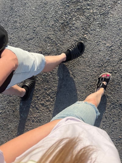 The image shows a top-down view of two people's legs walking on an asphalt surface. The person on the left is wearing light blue denim shorts and black slide sandals with black socks. The person on the right is wearing similar light blue denim shorts and black platform sandals with buckles, revealing bright pink toenails. Both individuals appear to be in motion, casting shadows on the textured gray asphalt. The lighting suggests it is daytime, likely outdoors. The environment is a paved surface, consistent with a road or walkway. There is no visible indication of the location being specifically within the city of Mal'tsevo, Russia, or any other identifiable urban area.