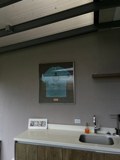 The image shows a kitchen in San Rafael, Costa Rica, located at the coordinates 9.966, -84.181. The kitchen features a white countertop with a stainless steel sink and a silver faucet. A small framed picture of a cartoon drawing is sitting on the counter. A framed, blue, framed shirt with the words “Jeep” and “1st Place” is hanging on the gray wall.  There is a wooden shelf with a decorative object on it, located to the right of the framed shirt. The kitchen has a wooden cabinet and a partial view of the ceiling with grey beams.