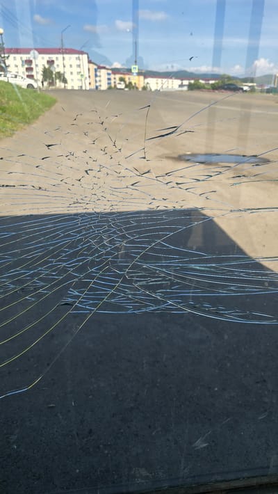 The image depicts a cracked windshield, likely from a vehicle, overlooking a street scene in Shakhtyorsk, Russia. The windshield is extensively fractured with a web of fine cracks radiating outwards. Through the damaged glass, a street is visible, with buildings lining one side and a grassy embankment on the other. The sky is blue with scattered clouds, and the sun casts a shadow on the asphalt below, suggesting it is daytime. Several cars are parked or driving on the street, and there are streetlights and a pedestrian crossing sign visible. The buildings appear to be apartment blocks with red roofs. The overall scene conveys a sense of everyday life in a Russian urban environment, with the damaged windshield serving as a prominent focal point.