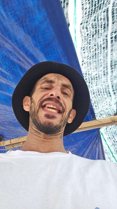 An adult man is captured in a close-up portrait, smiling broadly with an open mouth, showing his teeth, and looking directly at the camera. He wears a dark bucket hat and a white t-shirt, with part of his upper body visible. He has a prominent beard and several tattoos on his face, including on his forehead and cheekbones.

The man appears to be outdoors, situated under a blue tarp or fabric canopy that stretches across the top and right side of the frame, providing shade. Light brown bamboo poles are visible in the background, supporting the overhead structure. Below the blue tarp, a textured material with a white and black patterned mesh or netting is also visible. The bright lighting suggests it is daytime. The man's expression is jovial and happy. No visible text is present. The scene is set in Bac Cam Ranh, Vietnam.