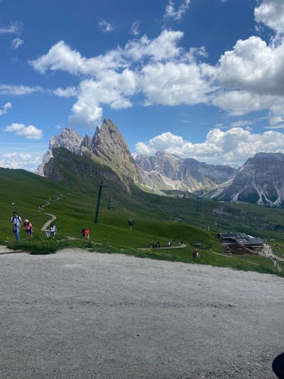 The image depicts a panoramic view of a mountain landscape in Santa Cristina Gherdëina, Italy, under a partly cloudy blue sky. In the foreground, a gravel path leads the eye towards a vibrant green meadow dotted with yellow wildflowers. Several individuals and groups are seen walking along a winding path that traverses the meadow, some descending and others ascending the gentle slope. A cable car support tower stands prominently on the left side of the path.
In the mid-ground, a cluster of wooden buildings, resembling a mountain lodge or restaurant, is situated. Construction activity is visible near one of the buildings, with heavy machinery present. The path continues towards these structures, with more people milling about.
The background is dominated by dramatic, jagged mountain peaks, characteristic of the Dolomites. The imposing rock formations rise sharply against the sky, their surfaces showing layers and crevices. The distant mountains are shrouded in a hazy, atmospheric perspective, indicating their significant distance. The overall scene suggests a popular tourist destination, likely during the daytime and possibly summer, given the green vegetation and the attire of the people.