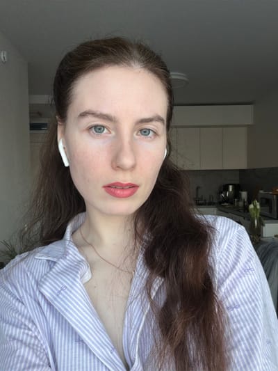 This image captures a chest-up portrait of an adult female in an indoor residential setting, specified as Ковалёво, Russia. The individual, with fair skin, blue-green eyes, and long, dark brown hair styled with a central part, is looking directly at the camera with a neutral expression. She is wearing two white wireless earbuds and has a light pink or coral lipstick applied. Her attire consists of a light purple and white striped collared shirt, partially unbuttoned, revealing a thin rose gold-colored necklace. Small purple bow motifs are visible on her shirt. The background to the viewer's left features a light-colored wall with a partial view of a green potted plant. To the viewer's right, a modern kitchen area is discernible, equipped with light-colored upper cabinets, a dark countertop, a stainless steel stand mixer, a sink, a vase containing white flowers (likely tulips), and a portion of a microwave oven. A circular ceiling light fixture is visible in the upper background. Natural light appears to illuminate the scene from the left.