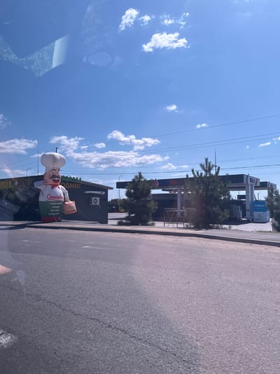 In Usatove, Ukraine, on a bright, sunny day with a clear blue sky dotted with white clouds, a scene unfolds along a paved road. To the left, a large inflatable chef, standing approximately 15-20 feet tall, is positioned in front of a dark-colored building with a sign bearing Cyrillic lettering. The chef, dressed in a white chef's hat and outfit, holds a brown box and appears to be waving. The building behind the inflatable figure has an air conditioning unit mounted on its exterior wall. Adjacent to this building, and further to the right, is a "FLY OIL" gas station with pumps and signage. Several evergreen trees are scattered throughout the scene, partially obscuring the view of the gas station. The foreground consists of the asphalt road, with a concrete curb and a sidewalk area visible. The overall impression is that of a commercial area on the outskirts of town.
