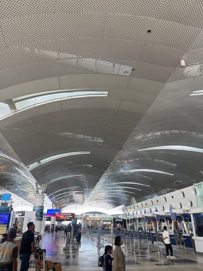 The image shows the interior of Kuala Namu International Airport, located in Ramunia Dua, Indonesia. The perspective is lengthwise through a curved tunnel-like walkway. The curved walls and ceiling are constructed of panels with a dotted pattern and long window-like openings. Along the right side of the walkway, there are airline check-in counters with digital displays. People are standing at the counters, presumably checking in for flights. Others are walking through the tunnel with luggage. The floor is glossy and reflects the overhead lights and signage. The lighting is bright and appears to be natural light coming through the window openings in the ceiling and walls.
