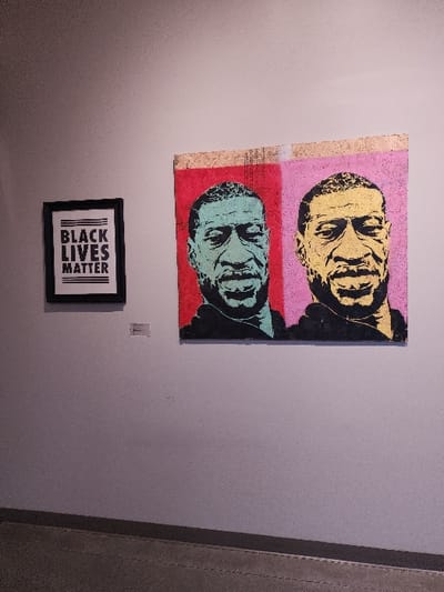 The image depicts an interior view of an art exhibition, likely situated within the Northwest African American Museum (NAAM) in Seattle, Washington, United States. A plain light grey wall displays two prominent artworks. On the right, a large rectangular painting in a pop-art style features two side-by-side portraits of George Floyd. The left portrait shows a teal-colored face against a red background, while the right portrait displays a gold-colored face against a pink background, both rendered with dark outlines. To the left of this painting, a smaller, framed black sign with white text reads "BLACK LIVES MATTER" in a bold, sans-serif font. A small, unreadable informational label is affixed to the wall below the "BLACK LIVES MATTER" sign. The floor is a darker grey, possibly concrete, exhibiting subtle linear patterns. No individuals are present in the scene.