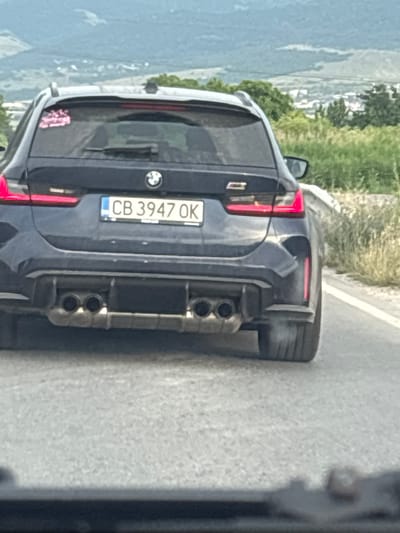 The image shows the rear of a dark-colored BMW wagon with Bulgarian license plates reading "CB 3947 OK". The car is on a paved road, with a grassy shoulder and trees in the background. A town or village is visible in the distance, nestled in a valley with hills beyond. The sky is overcast, and the overall lighting suggests it is daytime. There are no people clearly visible in the image. The car's exhaust pipes are visible and appear to be emitting some smoke or steam. The scene is likely outside of the town of Yoakim Gruevo, Bulgaria, given the landscape and the Bulgarian license plate.
