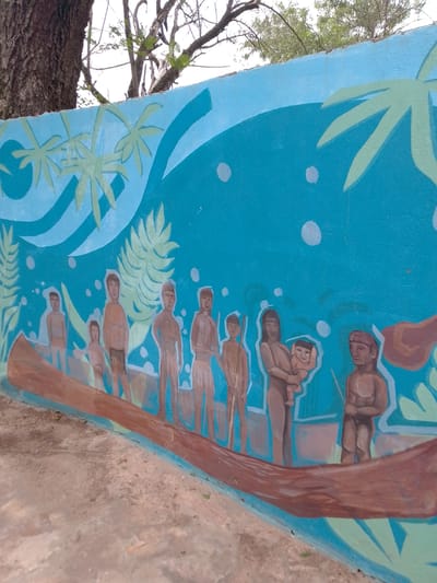 The image shows a mural in Villa Hayes, Paraguay. The mural depicts indigenous people in a boat, set against a background of blue and green with palm trees and circular shapes. The people in the boat are rendered in a primitive style, with several adults and one child. One adult figure holds a baby. The boat itself is brown and spans the length of the mural. The mural is painted on a concrete wall or barrier, and a portion of a tree is visible adjacent to the wall. The ground in front of the wall appears to be concrete.