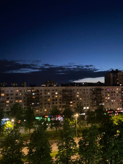 The image shows a night scene of Vaganovo Massiv, Russia. The left side of the image is dominated by a dark blue sky with a few clouds visible near the horizon. A tall, prefabricated apartment building occupies the right side of the frame, its many windows lit up with warm, yellow light. Trees line the bottom right edge of the building, partially obscuring the facade. A street with visible lane markings runs alongside the trees, illuminated by streetlights. The building's facade is plain and utilitarian, typical of Soviet-era architecture. A small number of colorful neon lights can be seen on the higher floors of the building on the right, presumably a business. The image appears to have been taken from a relatively low angle.