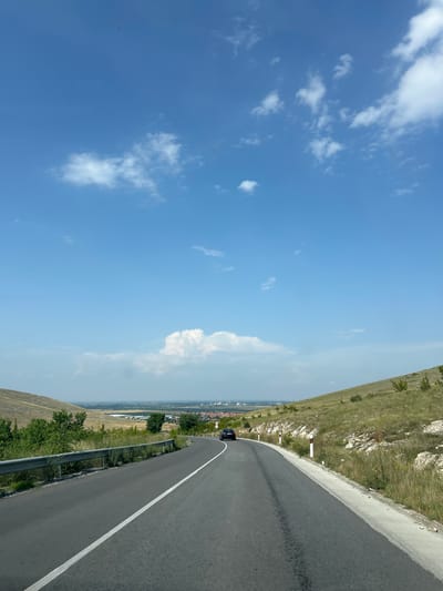 The image displays a clear daytime scene of a paved road winding through a hilly, semi-arid landscape near Isperihovo, Bulgaria. The road, dark asphalt with a solid white line on its left edge, curves right in the foreground and continues into the distance. A single dark-colored sedan is visible on the right side of the road, traveling away from the viewer. To the left of the road, a silver metal guardrail runs alongside a vegetated embankment of green bushes and dry, yellowish-brown grass leading up a hill. To the right, white and red reflective roadside markers delineate the edge of the road, followed by a concrete curb and then a rocky, grassy slope covered in dry vegetation and sparse small trees. In the middle ground, through the valley framed by the hills, a settlement identified as Isperihovo is visible, characterized by numerous buildings with red roofs. Beyond the settlement, industrial structures, possibly chimneys, and flat agricultural fields are discernible under a hazy sky. The sky above is a bright blue with scattered white cumulus clouds, particularly clustered near the horizon. The scene is brightly illuminated by natural sunlight, casting visible shadows.