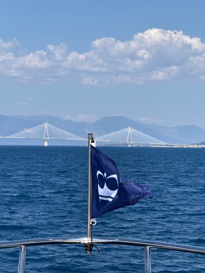 The image shows a blue flag with a white logo on a boat moving through the ocean. The flag is positioned on the right side of the frame, with a metal railing to its right. The background features a clear blue sky with scattered white clouds on the left and a vast expanse of deep blue water on the right. In the distance, a large suspension bridge is visible, spanning across the water. The bridge has tall white towers and a network of cables, suggesting it is a significant architectural structure. The image appears to be taken during the daytime, with bright sunlight illuminating the scene. The weather seems pleasant, with a clear sky and calm waters. The overall mood is serene and expansive, evoking a sense of travel and exploration. The logo on the flag is a stylized white symbol that resembles a bird or an abstract design, though its exact meaning is not discernible. There is no visible text in the image. The presence of the bridge and the type of flag suggest a location in Greece, specifically near Patras, which is known for the Rio-Antirrio Bridge.