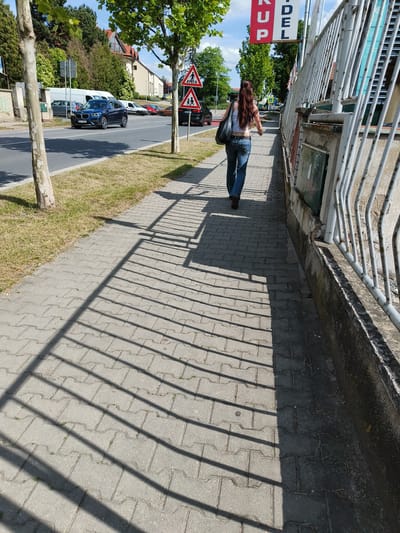 The image captures a sunny outdoor scene along a sidewalk next to a road, likely in Jesenice, Czechia. In the foreground, a woman with long, dark hair tied in a ponytail is walking away from the camera. She is wearing blue jeans, a white tank top, and brown shoes, with a dark shoulder bag on her left side. Her shadow, along with the distinct striped shadows cast by a fence, stretches across the paved sidewalk.

To the right, a white metal fence runs parallel to the sidewalk, separating it from a concrete wall and possibly a building. Above the fence, part of a red and white sign with the visible text "KUP" and "DEL" (likely part of a larger word like "PRODEJ" or "VÝKUP" related to commerce) is seen against a bright blue sky with some clouds.

To the left of the sidewalk, a narrow strip of dry grass separates it from a paved road. Several trees with light-colored bark stand along this strip, casting shadows. On the road, a blue SUV with a Czech license plate "FL4 00E" is visible, appearing to be in motion or parked. Other cars are also present on the road, including a white van and a red car further down. In the background, beyond the road, are residential or commercial buildings and dense green foliage.

Two red-bordered triangular warning signs depicting a walking person (pedestrian crossing) and two children (children playing or school zone) are positioned on a pole near the right edge of the road. The bright sunlight suggests it is mid-day or early afternoon. The overall scene depicts a typical street environment with pedestrian and vehicular traffic.