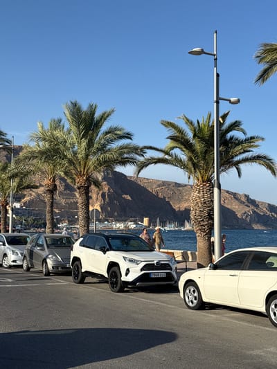 A photograph shows a waterfront promenade in Aguadulce, Spain, on a sunny day. Several palm trees are positioned along the promenade, with a light pole featuring two lamps also visible. In the background, a range of rocky hills forms the coastline, and the blue sea stretches out towards the horizon, dotted with sailboats.
In the foreground, a row of cars is parked on the asphalt. The closest vehicle on the right is a white sedan. To its left, a white SUV is parked, displaying the license plate "7946 LLC". Further to the left are two silver cars.
Several people are present on the promenade. Two individuals are walking towards the left, with one wearing a light-colored hat and shirt, and the other a patterned top. A person in pink shorts is standing near the white SUV. Another person in a patterned shirt and another in a light-colored hat are walking together in the distance, near the palm trees and the sea. The scene is captured from a ground-level perspective.