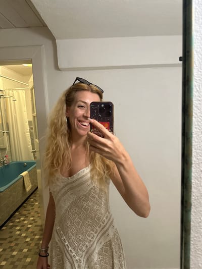 A blonde woman with light skin and fair complexion is taking a selfie in a bathroom. She is wearing a white lace dress, sunglasses on her head, and bracelets on her left arm. The woman is smiling widely and holding up a smartphone with a glittery case. Behind her, a turquoise bathtub and a tiled floor are visible. The bathroom appears to be in an older building, possibly in the city of Ammeldingen bei Neuerburg, Germany.