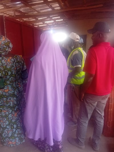 The image depicts a group of people gathered in what appears to be a sheltered, semi-enclosed outdoor space in Okengwe - Obehira, Nigeria.
On the far left, a person is seen from behind, wearing a colorful, patterned dress, likely made from local Ankara fabric, with a matching headwrap. Adjacent to them, another individual, also viewed from the back, wears a long, flowing light purple garment (possibly a jilbab or abaya) over a dark patterned dress. A bright light source creates a significant glare directly behind their head.
To the right, a man is visible wearing a white cap and a high-visibility vest in lime green or yellow, suggesting he might be involved in construction, safety, or an organized activity. Another man in a dark cap is partially obscured. On the far right, a man is seen from the back, wearing a dark baseball cap, a bright red short-sleeved polo shirt, and grey trousers.
The setting features an overhead structure, possibly a simple roof or canopy made of corrugated metal or planks. In the background, there is a reddish-brown wall or structure, and what appear to be stacked materials, possibly bricks. The people seem to be standing and interacting casually, perhaps conversing or waiting for something. The attire of the individuals, particularly the patterned fabrics, is consistent with the cultural context of Nigeria. There is no visible text in the image.