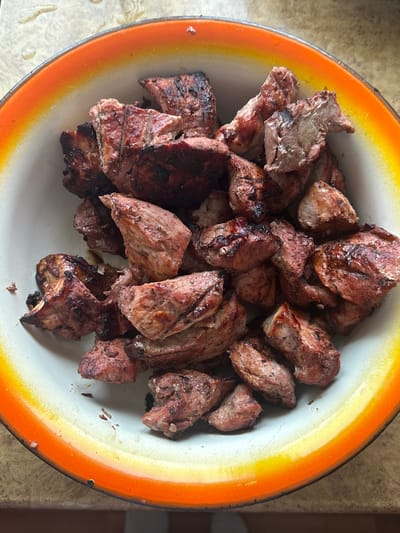 A top-down shot shows a bowl filled with pieces of cooked meat. The bowl has an orange rim and a white interior. The meat appears to be roasted or grilled, with some pieces showing darker char marks. The pieces are irregular in shape and size. The background is a neutral, slightly textured surface. The city and location are not discernible from the image.
