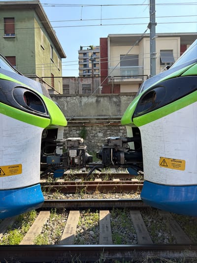 This media file depicts a close-up view of two modern trains, one green and white, the other white and blue, positioned side-by-side on train tracks in Lecco, Italy. The trains are stationary, suggesting they are either stopped at a station or in a yard. The foreground is dominated by the trains, with their sleek, aerodynamic designs visible. The upper portion of the image shows the front of the green and white train, and the lower portion shows the front of the white and blue train.
Between the trains, the mechanical components connecting them are visible, including intricate wiring and hydraulic systems. The tracks themselves are laid on ballast, with overgrown grass and weeds suggesting the location may be somewhat rural or less maintained. In the background, a residential building with multiple stories and balconies can be seen, along with overhead power lines for the trains. The sky is clear and blue, indicating a sunny day.
Two identical yellow warning stickers with black text are visible on the sides of the trains. The text, in Italian, reads "ATTENZIONE DURANTE L'ACCOSTO VIETATO INTRODURSI TRA I VEICOLI," which translates to "ATTENTION DURING DOCKING, DO NOT ENTER BETWEEN VEHICLES." This highlights a safety precaution related to the trains being coupled or uncoupled. The overall impression is one of industrial infrastructure in an urban or semi-urban setting on a clear day. There are no people visible in the frame.