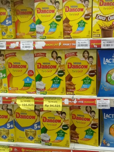 This image depicts a brightly lit interior view of a retail environment, likely a supermarket or grocery store, located in Nganjuk, Indonesia. The primary focus is a display of numerous "Nestle Dancow FortiGro" powdered milk products arranged on three white shelves.
The shelves are uniformly stocked with yellow rectangular cartons of various "Dancow FortiGro" variants, including "Full Cream," "Instant," and "Cokelat" (chocolate). Each carton features the "Nestle Dancow FortiGro" logo prominently displayed within a red heart shape, often accompanied by illustrations of smiling adults and children, glasses of milk, or chocolate beverages. Specific product weights, such as 390g, 750g, and 780g, are visible on some packages.
Price tags, predominantly white with black text, are affixed to the front edge of the shelves, indicating prices in Indonesian Rupiah (Rp). Examples include Rp 53,995, Rp 46,450, Rp 94,525, Rp 104,925, and Rp 90,025. Some price tags are overlaid with yellow "PROMO SANGAT MURAH" (very cheap promo) stickers. Orange shelf strips with white text, partially obscured, are also present, with visible phrases such as "ANAK 1 TAHUN" (1 year old child).
To the far right, blue cartons of "Lactamil" powdered milk products are partially visible on the top and bottom shelves. No individuals are present or actively interacting with the products in the immediate scene. The overall lighting is bright and even, consistent with commercial retail spaces.