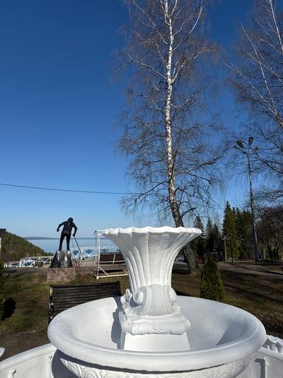 A low-angle view captures a stark contrast of urban elements against a natural backdrop, potentially in Galevo, Russia. A white, ornate vase-like structure dominates the foreground, positioned to the right. To the left, a skeletal birch tree reaches towards a cloudless blue sky. In the middle ground, a dark sculpture of a figure pulling something, possibly a skier, stands on a stone pedestal. A metal railing separates the figure from a body of water with a distant shoreline. Beyond the sculpture, a bench sits adjacent to the vase, with a backdrop of trees and a lamppost. The scene suggests a public park or monument area.