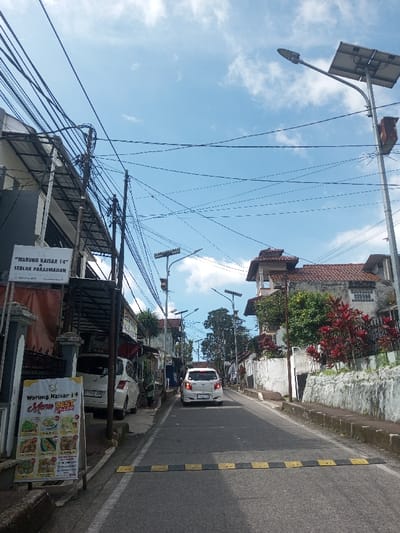 This image depicts an uphill residential street scene in the city of Naung Bo Oe, Myanmar, under a clear blue sky with scattered white clouds. In the immediate foreground, a black and yellow striped speed bump spans the width of the asphalt road. To the left, a white A-frame sign displays "Warung Kaisar 14" and "Menu Best Seller!" along with various food images. Behind it, a rectangular white sign is mounted, identifying "WARUNG KAISAR 14 & SEBLAK PARASMANAN" with contact information. Overhead, a dense network of black electrical and utility wires crisscrosses the sky, suspended from multiple dark utility poles that line the left side of the street.

Further up the left side, two white cars are parked along the curb, facing away from the viewer. A person, possibly wearing a green garment, is partially visible on the sidewalk beside the parked vehicles. Along the right side of the street, a white car, identified by its Indonesian license plate "B 1269 FZB", proceeds uphill. Numerous streetlights, each equipped with a solar panel, are installed on poles along both sides of the road, extending into the distance. The right side of the street features a low white wall adorned with red-leaved plants and green shrubs, behind which a multi-story building with a distinct red tiled roof and an ornate upper structure is visible. The street continues to ascend, flanked by residential structures and vegetation under daylight conditions.
