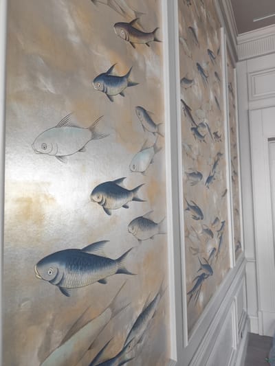 A close-up, slightly angled shot displays three panels adorned with fish artwork. The panels are framed with white molding and hang in a vertical series. The fish are depicted in various sizes and colors, with some appearing to be more detailed than others. The background of the panels features an abstract pattern with a mix of silver and gold hues. To the right of the panels is a white wall with a white door that has decorative molding. The setting suggests an interior space, possibly a hallway or corridor. The location is Форосский поселковый совет, Ukraine.