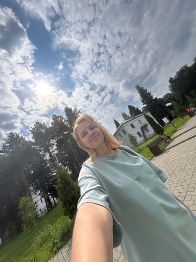 A young woman with blonde hair smiles at the camera, taking a selfie outdoors. She is wearing a light blue t-shirt and is partially visible from the chest up. The background reveals a sunny day with a bright sun and scattered clouds in a blue sky. To the left, tall green trees form a dense forest. In the distance, a white building with windows and a fire escape stands amidst greenery. A paved walkway, made of interlocking bricks, leads past benches and lampposts. The scene appears to be a park or recreational area in Breytovo, Russia.