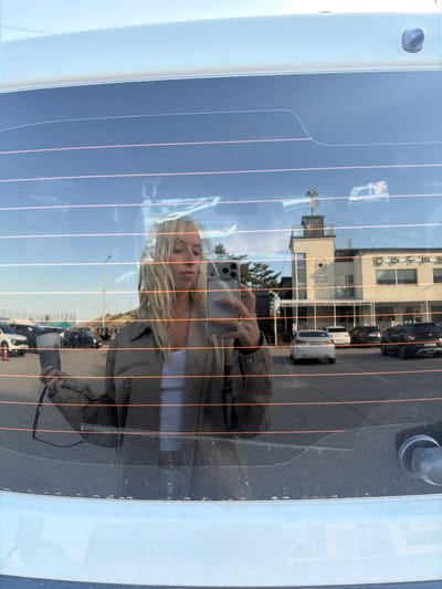 A reflective shot through the rear window of a white vehicle. The reflection shows a blond woman holding a phone to take a picture of herself. Outside the window, a building is visible with writing on it, alongside passing vehicles. The location is Toseong-myeon, South Korea.