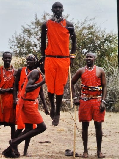 This image, taken in Ihumwa, Tanzania, displays five men outdoors. The central figure is suspended mid-air, performing a vertical jump, barefoot, and wearing a bright orange-red shuka belted at the waist, adorned with multiple white and blue beaded necklaces and wristbands. To his immediate left, another man in a similar red shuka with beaded accessories leans forward, holding a thin wooden stick. Further left, two additional men stand, also in red shukas and beaded jewelry; the leftmost man smiles. To the right of the central jumping figure, a fifth man stands, wearing a striped red shuka with a multi-layered beaded belt and necklaces, holding a light-colored wooden staff in his right hand while wearing dark sandals. A pair of dark sandals rests on the dry, light brown earth foreground near the staff. The background consists of sparse, dry bushes and trees under an overcast, light grey sky.