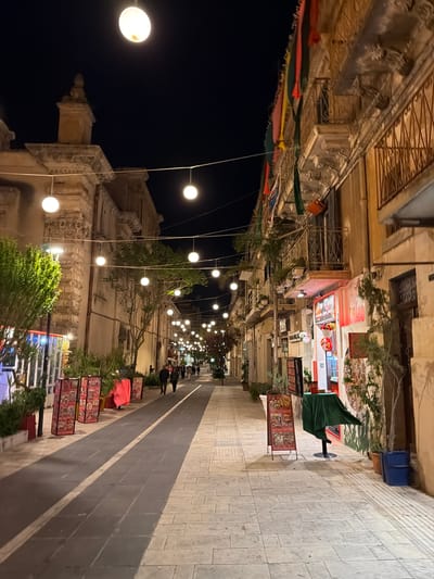 This image depicts a street scene at night in Ragusa, Italy. The street is lined with buildings, illuminated by hanging string lights. Red, yellow, and green flags are hung across the buildings. Several people are walking down the street. Along the right side of the street are food stands and displays. The street is paved with light-colored stones. A tall building is visible in the background on the left.