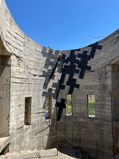 The image depicts the interior of a concrete structure located in Gornji Katun, Montenegro. The structure appears to be an abandoned building with exposed concrete walls and a curved archway. The walls feature numerous square or rectangular openings, potentially former windows, with glimpses of greenery visible through some. A pattern of large, black, cross-shaped graffiti is visible on the curved wall. Exposed rebar protrudes from the concrete in various places. The sky is visible through the archway.
