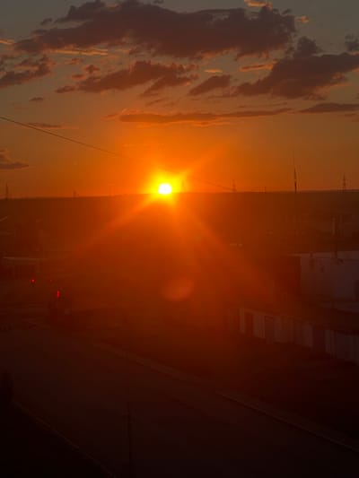 This is a vertical, wide-angle photograph of a sunset over an urban landscape, possibly Satbayev, Kazakhstan. The sun, a brilliant orb of intense orange light, is the dominant subject, positioned in the left half of the frame. It casts long rays that create a lens flare effect and illuminate the surrounding sky in warm hues of orange and yellow. Wispy, pink-tinged clouds are visible in the upper left, suggesting a picturesque sunset.
The scene transitions from the bright sky to a darker, silhouetted cityscape. Tall structures, possibly buildings or industrial elements, are visible against the sunset. Power lines or cables stretch across the frame, crossing the sky and some of the structures, adding a sense of depth and human infrastructure to the natural spectacle. In the mid-ground, lights on a structure, appearing as small red dots, suggest activity within the urban environment. The lower right portion of the image is predominantly dark, with faint lines that might indicate streets or architectural features.
The time of day is clearly sunset, characterized by the low angle of the sun and the warm, saturated colors of the sky. The weather appears clear enough for a vibrant sunset, with clouds adding texture. There is no discernible text in the image. The overall mood is one of natural beauty juxtaposed with an industrial or urban setting, evoking a sense of calm and perhaps introspection as day turns to night.