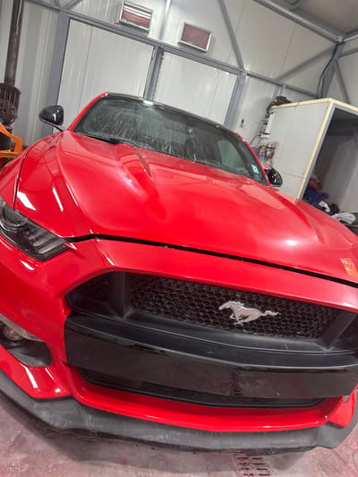 The image shows a red Ford Mustang parked inside a garage or workshop. The car is prominently positioned in the foreground, with its front end facing the viewer. The grill features a silver Mustang emblem. The car appears to be indoors, possibly undergoing maintenance or repairs, given the setting. The garage is constructed with white panel walls and a metal framework. The location is presumed to be Golyanovtsi, Bulgaria. There are no people visible in the image.